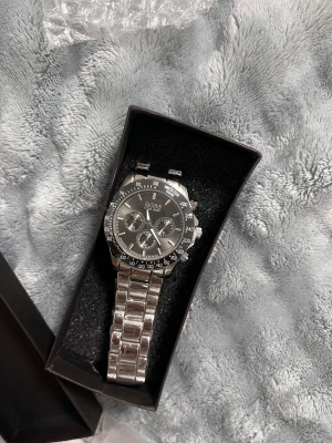 Hugo Boss silverfärgad klocka - Stilren klocka från Hugo Boss med silverfärgat armband i metall och svart urtavla. Klockan har tre mindre urtavlor, tydliga index och detaljer i guldton. Länken är robust och låset är graverat med BOSS-loggan. Perfekt accessoar för dig som gillar klassisk design.