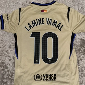 Barcelona Lamine Yamal 10 Nike Dri-FIT tröja - Snygg Barcelona fotbollströja i beige med svarta detaljer, Lamine Yamal 10 på ryggen. Nike Dri-FIT material som andas, klubbmärke och Spotify-logga framtill. Blå och svarta ränder vid krage och ärmslut. Perfekt för dig som älskar Barca och vill sticka ut.