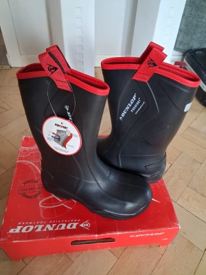 Svarta Dunlop Purofort stövlar med röd kant - Säljer ett par svarta Dunlop Purofort stövlar med röd kant och grov sula i svart och rött. Stövlarna har praktiska dragflikar upptill och är tillverkade i slitstark syntet med isolerande egenskaper. Perfekt för blöta och kalla dagar, riktigt robusta och snygga... ny pris 1800 kr