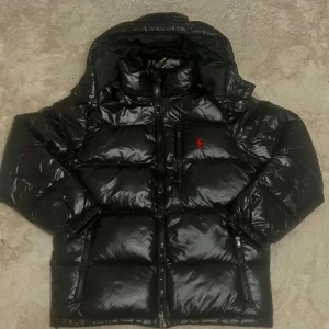 Svart pufferjacka från Polo Ralph Lauren - Säljer en svart pufferjacka från Polo Ralph Lauren med glansig finish och röd logga på bröstet. Jackan har huva, flera dragkedjefickor och tvåvägsdragkedja framtill. Perfekt för kalla dagar och har en riktigt schysst streetstil.