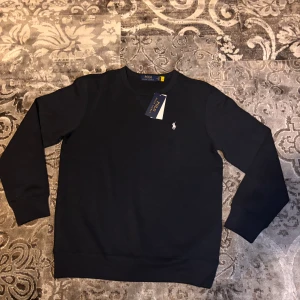 Svart  Polo Ralph Lauren sweatshirt - Svart sweatshirt från Polo Ralph Lauren med klassisk rund hals och broderad vit logga på bröstet. Tröjan har långa ärmar och ribbade muddar vid ärmslut och nederkant. Perfekt för en clean och stilren look.