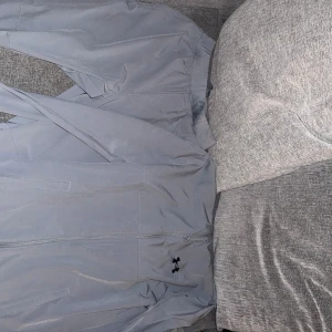 Grå vindjacka från Under Armour - Snygg grå vindjacka från Under Armour med dragkedja framtill och diskret logga på bröstet. Jackan har långärmade ärmar och är tillverkad i lätt polyester som passar perfekt för träning eller chill. Enkel och stilren design som funkar till många outfits.