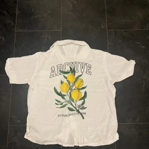 Vit skjorta med citronprint från Solid - Vit skjorta från Solid med coolt citrontryck och texten 'ARCHIVE' på ryggen. Framtill finns en bröstficka med samma print. Skjortan har korta ärmar, klassisk krage och knäppning framtill. Perfekt för dig som gillar fräscha och unika detaljer.