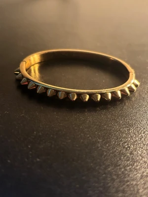 Guldarmband med nitar från Edblad - Snyggt guldarmband från Edblad med coola konformade nitar runt hela bandet. Armbandet är stelt och har en modern, edgy vibe som verkligen sticker ut. Perfekt för dig som gillar accessoarer med attityd och vill ha något som syns.