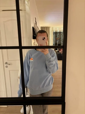 Ljusblå sweatshirt från Ellesse - Säljer en ljusblå sweatshirt från Ellesse med klassisk logga i orange och rött på bröstet. Tröjan har rund hals, ribbade muddar och är tillverkad i mjuk bomull och polyester.