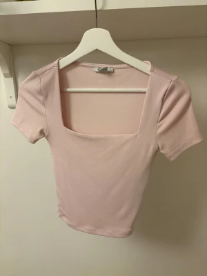Ljusrosa kort topp med fyrkantig hals - Säljer en ljusrosa topp med korta ärmar och snygg fyrkantig halsringning. Toppen är enkel och stilren, perfekt att matcha med jeans eller kjol. Materialet känns mjukt och stretchigt, vilket gör den bekväm att bära hela dagen. Tröjan är tjockare i materialet framtill så den ger mer täckning där. 😊
