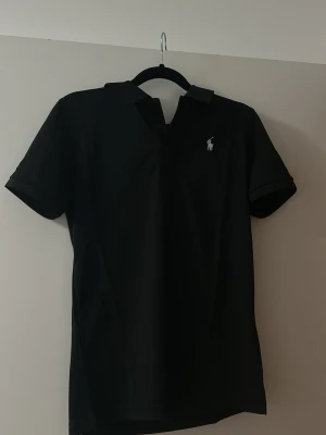 Svart piké från Polo Ralph Lauren - Svart klassisk pikétröja från Polo Ralph Lauren med vit broderad logga på bröstet. Tröjan har krage, korta ärmar och knappar vid halsen. Materialet är mjuk bomull som ger en skön känsla och snygg passform. Perfekt för dig som gillar stilrena och tidlösa plagg. Storlek S