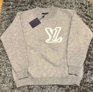 Grå stickad Louis Vuitton tröja - Snygg grå stickad tröja från Louis Vuitton med stort vitt LV-logo framtill. Tröjan har rund hals, ribbade muddar och är långärmad. Perfekt för dig som gillar exklusiva märken och vill ha en stilren look.