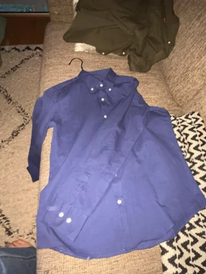 Blå skjorta från Polo Ralph Lauren - Snygg blå skjorta från Polo Ralph Lauren med klassisk krage och vita knappar. På bröstet finns den ikoniska röda Polo-loggan broderad. Skjortan är långärmad och gjord i mjuk bomull, perfekt för dig som gillar stilrena och tidlösa plagg. pris är inte hugget i Sten