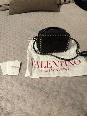 Svart axelväska Valentino Garavani - Säljer en svart axelväska från Valentino Garavani med fyrkantig form och coola guldfärgade nitar runt kanterna. Väskan har justerbar axelrem med nitar och dragkedja upptill. Materialet är äkta skinn och loggan syns i guld på framsidan. Dustbag och äkthetskort medföljer. Köptes för 2 år sedan för ca 14000 men säljer den nu för 4000 (kan diskutera pris). Den är mycket sparsamt använd och har inte en endaste skråma💕💕💕💥💥💥