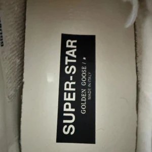 Golden Goose Superstar sneakers vit/grå - Golden Goose Superstar sneakers i vitt skinn med grå mockadetaljer och snörning. Ikonisk grå stjärna på sidan och textdetaljer i guld. Platt sula och rund tå. Snyggt slitna detaljer för en cool look. Perfekt för dig som gillar italiensk design och streetstyle. Säljer dem pga av att dem är stora på mig. Passar som 43 också. Aldrig använt, helt nya.  