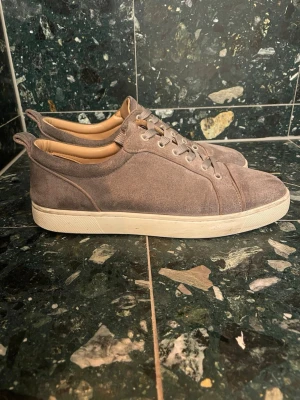 Grå Christian Louboutin  - Snygga grå loubs med vit platt sula och coolt röd undersida. Skorna har rund tå, snörning och ventilerande hål på sidan. Insidan är beige och de har en stilren, minimalistisk look som passar alla. Saknar en knapp på sidan. Storlek 42
