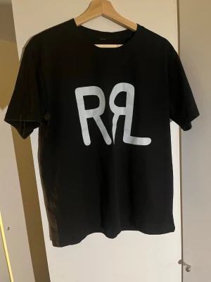 RalphLauren T-shirt - Svart t-shirt med stor vit RL-logga på bröstet. Klassisk rund hals och korta ärmar. Perfekt för dig som gillar enkel stil med tydlig logga.