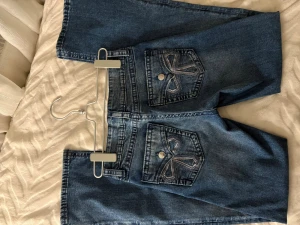 Blå jeans från Ginatricot med broderi - Snygga blå jeans från Gina Tricot i bootcut och coola broderade detaljer på bakfickorna. Klassisk femficksmodell med knapp och dragkedja framtill. Perfekt för dig som gillar att klä sig med stil! I barnstorlek men motsvarar ungefär 34 i vuxenstorlek! 