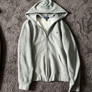 Grå hoodie från Polo Ralph Lauren - Snygg ljusgrå hoodie från Polo Ralph Lauren med dragkedja framtill och klassisk huva. Tröjan har två fickor, broderad logga på bröstet och är tillverkad i mjuk bomullsmix. Perfekt för chill dagar eller när du vill ha en clean look.