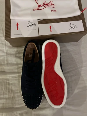 Christian Louboutin svarta sneakers strl 39 - Säljer ett par svarta sneakers från Christian Louboutin i storlek 39. Skorna har klassisk röd sula, snörning och coola svarta nitar på tån. Tillverkade i skinn och mocka för en lyxig känsla. Kommer med originalkartong och dustbag.