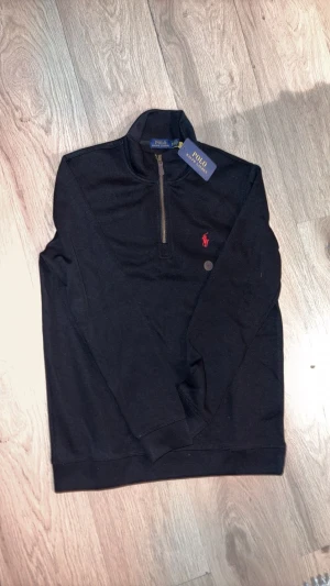 Svart half zip tröja från Polo Ralph Lauren - Svart långärmad tröja från Polo Ralph Lauren med half zip och ståkrage. Tröjan har det klassiska röda Polo-logot broderat på bröstet och är tillverkad i mjuk bomull. Perfekt för dig som gillar stilrena och tidlösa plagg.