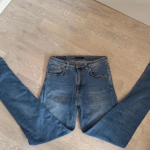 Blå skinny jeans från Nudie Jeans, w29 L32 - Snygga blå skinny jeans från Nudie Jeans med klassisk femficksdesign och orangea sömmar. Högmidjade med smal passform och stretchigt jeanstyg. Bakfickan har Nudie Jeans signaturbroderi och läderpatch. Perfekt för dig som gillar stilrena och bekväma jeans.