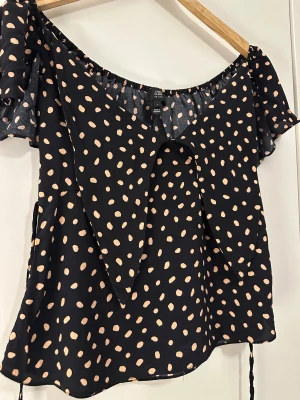 Svart prickig blus från River Island - Superfin svart blus med ljusa prickar från River Island. Modellen är offshoulder med korta, vida ärmar och stor krage framtill. Blusen har knytband i midjan och är gjord i ett lätt, flowy material som känns luftigt och skönt.
