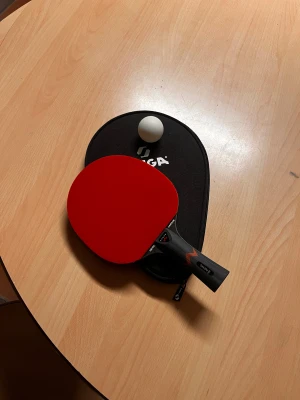 Svart och röd pingisväst från Stiga - Pingisväst från Stiga i svart och rött, med klassisk rund form och greppvänligt handtag. Västen har en slät svart sida och en röd sida med struktur, perfekt för att variera spelstil. Materialet är syntet och västen kommer med ett svart fodral med Stiga-logga. Samt kommer med en vit pingisboll till o spela med. Racket är väldigt bra och gör dig till en bättre spelare ”om man har bra och snygg utrustning spelar man ännu bättre”