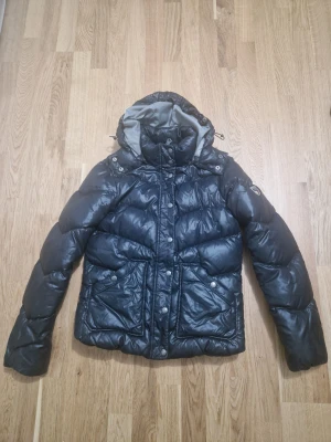 Mörkblå pufferjacka från Hilfiger Denim - Mörkblå pufferjacka från Hilfiger Denim med glansig finish och grått foder. Jackan har huva med dragsko, stora framfickor med tryckknappar och snygga quiltade detaljer. Perfekt för en modern, urban look.