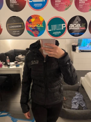 Svart jacka från EA7 Emporio Armani - Säljer en svart pufferjacka från EA7 Emporio Armani med huva och dragkedja framtill. Jackan har quiltad design och logga på bröstet. Perfekt för dig som vill ha en stilren och lätt jacka till höst och vinter.