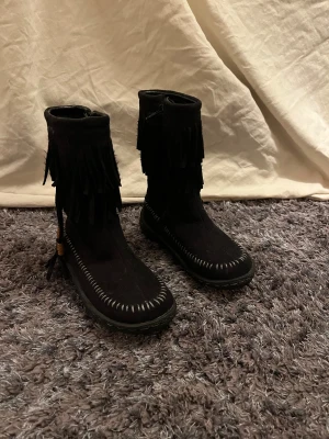 Svarta mockaboots med fransar från Duffy - Svarta boots i mocka från Duffy med snygga fransdetaljer runt skaftet och dekorativa träpärlor på sidan. Skorna har dragkedja på insidan och grov sula för extra grepp. Synliga vita sömmar längs tån ger en cool kontrast.