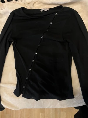 Svart långärmad topp med knappar - Säljer en svart långärmad topp från M&S med en unik diagonal rad av svarta knappar framtill. Toppen har en rund halsringning och är gjord i ett mjukt, ribbat material som sitter snyggt på kroppen. Perfekt för dig som gillar stilrena detaljer.