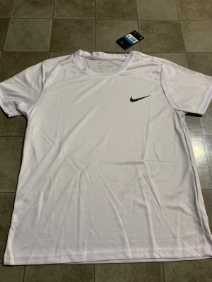 Vit Nike Dri-FIT t-shirt herr M - Fräsch vit t-shirt från Nike med svart swoosh-logga på bröstet. Tillverkad i lätt och ventilerande Dri-FIT polyester, perfekt för träning. Klassisk rund hals och korta ärmar. Snygg och sportig stil som passar dig som gillar att hålla dig aktiv.