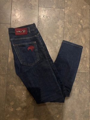 Stefano Ricci red eagle - Snygga mörkblå jeans från Stefano Ricci med exklusiva röda broderier på fickorna och ett vinrött märke med metallogo bak. Jeansen har klassisk femficksdesign, kontrastsömmar och är tillverkade i slitstarkt denim. Perfekt för dig som gillar detaljer och lyxig känsla. Nypris 12000 kr. Saknar alla etiketter. Bra skick. Säljer då de är försmå. 