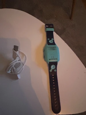 Barn GPS Smartklocka - Smartklocka för barn med GPS-funktion, blå/svart armband med lekfull design. Levereras med laddkabel. Bandet är lite slitet men är lätt att byta ut. Perfekt för föräldrar som vill hålla koll på barnets position.