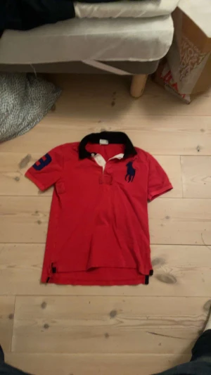 Röd Polo Ralph Lauren t-shirt - Snygg röd t-shirt från Polo Ralph Lauren med klassisk svart krage och blå logga på bröstet. Kortärmad modell med nummerdetalj på ärmen. Perfekt för dig som gillar sportig och stilren design. Materialet är mjuk bomull.
