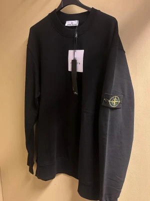 Svart sweatshirt från Stone Island - Svart sweatshirt från Stone Island med klassisk rund hals och ribbade muddar. Tröjan har lång ärm och den ikoniska Stone Island-loggan på vänster ärm. Tillverkad i mjuk bomull som är skön mot huden. Perfekt för dig som gillar stilrena och snygga streetwear-plagg.