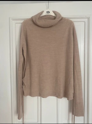 Beige polotröja med snörning Love Knitwear - Snygg beige polotröja från Love Knitwear med ribbad krage och långa ärmar. Tröjan har cool snörningsdetalj i sidan och är stickad i mjukt material. Perfekt för dig som gillar stilrena och trendiga plagg med en twist.