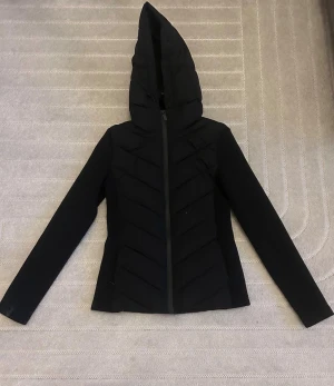 Svart pufferjacka med huva - Svart pufferjacka med stor, spetsig huva och dragkedja framtill. Jackan har quiltade partier på framsidan och långa ärmar. Snygg, stilren look med en smal passform och diskreta detaljer. Perfekt för kyliga dagar när du vill hålla dig varm och se cool ut.