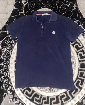 Mörkblå Moncler pikétröja i bomull - Snygg mörkblå pikétröja från Moncler med klassisk krage och korta ärmar. Tröjan har knappar framtill och Moncler-logga broderad på bröstet. Röd och vit randig detalj på insidan av kragen och vid knäppningen. Perfekt för dig som gillar stilrena märkesplagg.