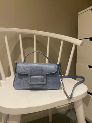 Ljusblå axelväska  - Snygg ljusblå axelväska från Zara med krokopräglat mönster och silverfärgad kedja. Väskan har ett stilrent spänne framtill, axelrem och ett rymligt fack med innerficka. 