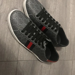 Gucci sneakers med GG-mönster - Säljer ett par snygga Gucci sneakers med klassiskt GG-mönster i grått och svart. Skorna har svarta snören, vit sula och de ikoniska blå-röda ränderna på sidan. Baktill är ena skon röd och den andra svart med metallic-finish och Gucci-logga. Perfekt för dig som gillar lyxiga detaljer.