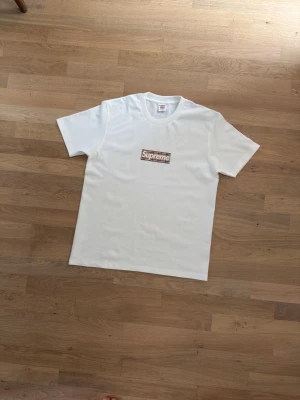 Supreme x Burberry t-shirt M - Vit t-shirt från Supreme x Burberry i storlek M. Klassisk rund hals och korta ärmar. Supreme-loggan på bröstet med Burberrys ikoniska rutiga mönster. Tillverkad i 100% bomull för skön och mjuk känsla. Perfekt för dig som gillar streetwear och designer.
