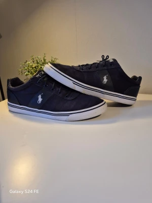 Mörkblå sneakers från Polo Ralph Lauren - Snygga mörkblå sneakers från Polo Ralph Lauren med vit logga på sidan. Skorna har snörning, vit sula med svarta ränder och är tillverkade i canvas och syntet med detaljer i skinn. Perfekta för en stilren och avslappnad look.