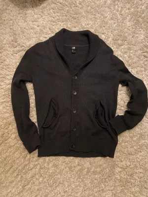 Svart kofta från H&M med knappar - Svart kofta från H&M med ribbad krage och muddar, knäppning framtill och två fickor med knappdetaljer. Långärmad med förstärkta armbågslappar i svart tyg. Perfekt för lager-på-lager och chill stil.