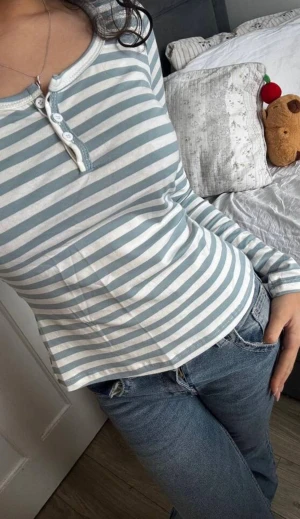 Randig vit blå långärmad topp  - Säljer en långärmad topp med ränder i ljusblått och vitt. Toppen har rund halsringning och en knappslå med fyra vita knappar framtill. Materialet är mjukt och stretchigt bomull. Fick den från en kompis så vet inte vart den är ifrån ser inte lappen men orginal priset va ca 130 sa hon🫶🏼