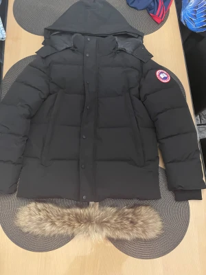 Svart Canada Goose dunjacka med päls - Säljer en svart dunjacka från Canada Goose med avtagbar huva och pälsdetalj. Jackan har ett klassiskt märke på ärmen, knappar framtill och två sidofickor. Perfekt för kalla dagar med sin tjocka, quiltade design och snygg look.