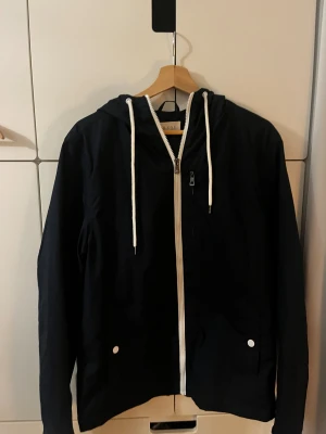 Svart vindjacka med huva och dragkedja - Svart vindjacka från pier one med vit dragkedja och vita snören i huvan. Jackan har två fickor med vita knappar och en extra bröstficka med dragkedja. Perfekt för dig som gillar stilren och enkel design med funktionella detaljer.