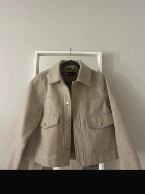 Beige cardiganjacka från ZARA - Stilren beige cardiganjacka från ZARA med klassisk krage och stora fickor framtill. Jackan har raka ärmar, tryckknappar och är tillverkad i ett mjukt, ull-liknande material. Perfekt för lager på lager och enkel att matcha med olika outfits.