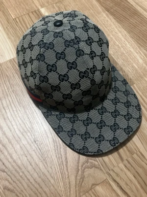 Gucci keps med GG-mönster - Snygg keps från Gucci i beige och svart med klassiskt GG-monogram över hela kepsen. Kepsen har böjd skärm och en röd-grön rand på sidan. Tillverkad i canvasmaterial med mörkblått innerfoder. Perfekt för dig som vill sticka ut med en lyxig accessoar.
