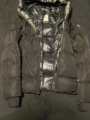 Svart dunjacka från Moncler - Snygg svart dunjacka från Moncler med stor huva och logotyptryck längs kanten. Jackan har två dragkedjefickor framtill, ribbade muddar och glansigt innerfoder. Perfekt för kalla dagar och riktigt trendig stil. Oops Kallebilderna är lite slitna men dem går och lagra. Den är användbar:)