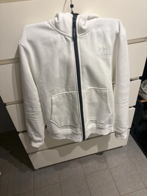 Vit hoodie från C.P. Company - Säljer en vit hoodie från C.P. Company med dragkedja framtill och två stora fickor. På bröstet finns en broderad logga och huvan har de ikoniska glasögon-linserna. Hoodien är i mjukt material och har en clean, modern look.