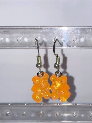 Orange godisnalle örhängen - Säljer ett par unika örhängen med orangea godisnalle-berlocker i plast. De har krokar i metall och en lekfull, trendig design som verkligen sticker ut. Perfekta för dig som gillar färgglada accessoarer och vill addera något kul till din stil.