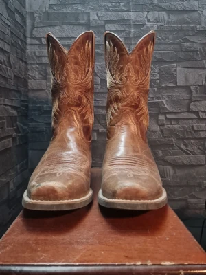Bruna klassiska cowboy boots - Snygga bruna cowboy boots i läder med dekorativa broderier längs skaftet. Bootsens spetsiga tå och robusta klack ger en autentisk westernkänsla. Dragflik på sidan för enkel påtagning. Perfekta för dig som vill ha en tidlös westernstil. Ny pris 2200